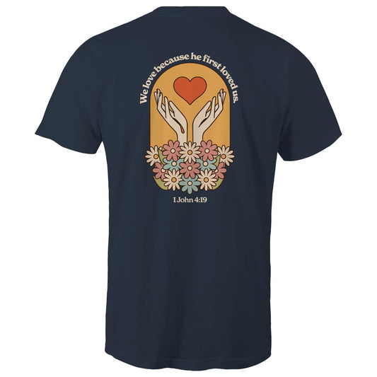 Christian t-shirt with bold faith-based design on the back featuring a red heart, outstretched hands, and colorful flowers. Includes the Bible verse “We love because he first loved us – 1 John 4:19.” Designed by Lighthouse Christian Clothing to inspire love and faith.