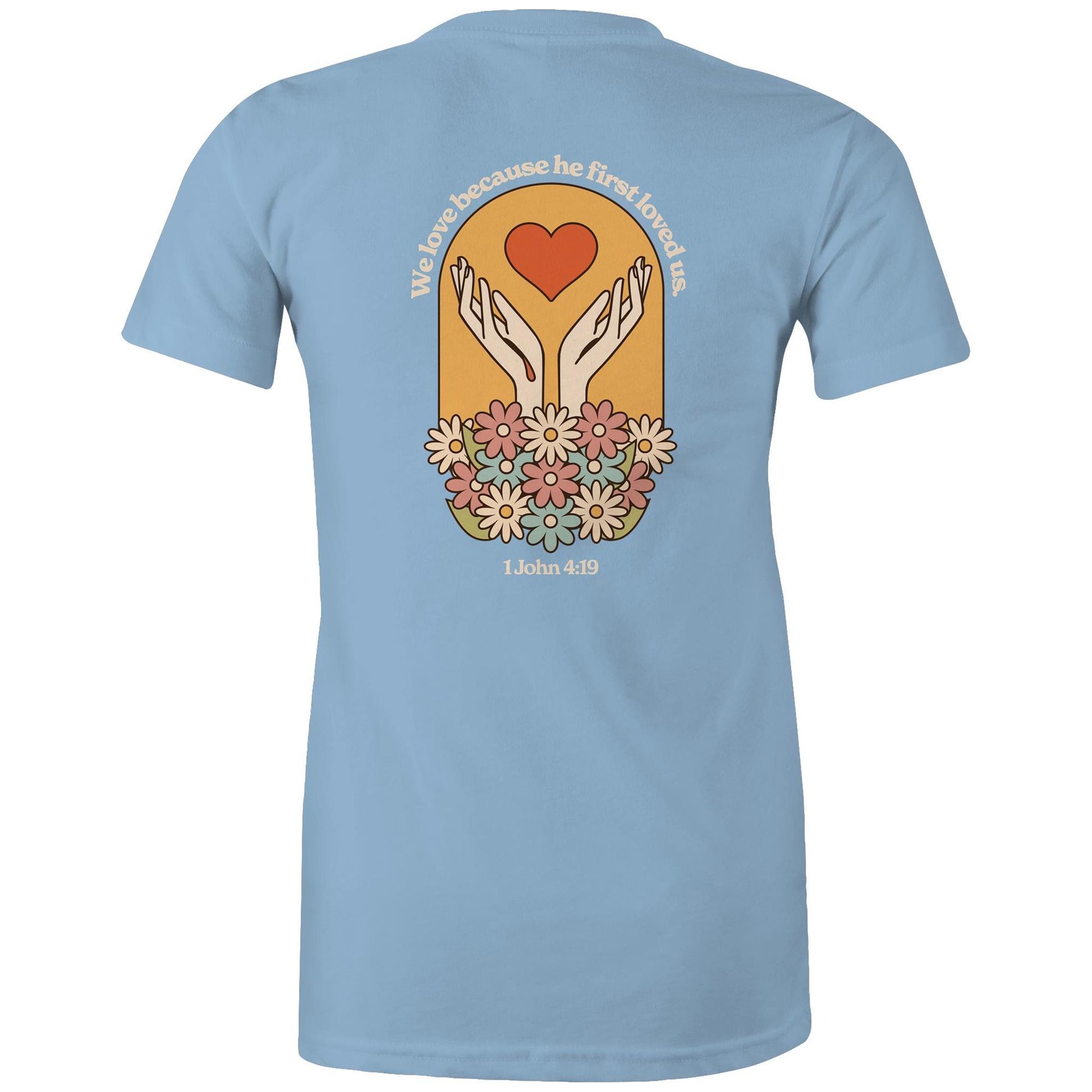 Christian t-shirt with bold faith-based design on the back featuring a red heart, outstretched hands, and colorful flowers. Includes the Bible verse “We love because he first loved us – 1 John 4:19.” Designed by Lighthouse Christian Clothing to inspire love and faith.