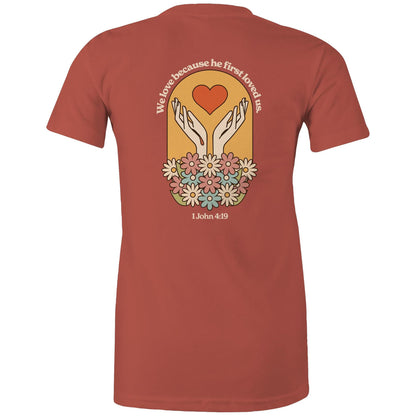 Christian t-shirt with bold faith-based design on the back featuring a red heart, outstretched hands, and colorful flowers. Includes the Bible verse “We love because he first loved us – 1 John 4:19.” Designed by Lighthouse Christian Clothing to inspire love and faith.